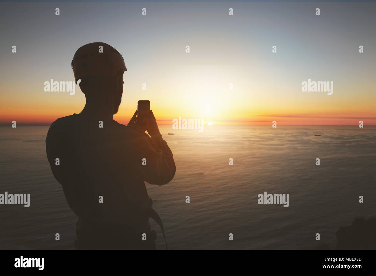 Silhouette rocciatore con la fotocamera del telefono tranquillo fotografare il tramonto sull'oceano Foto Stock