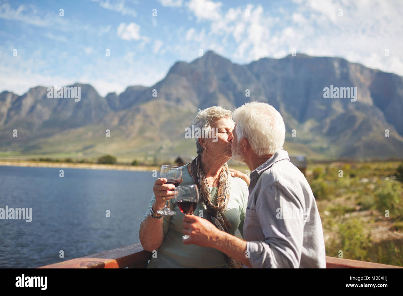 Romantico, attivo coppia senior baciare e un bicchiere di vino sul soleggiato balcone estivo a Lakeside Foto Stock