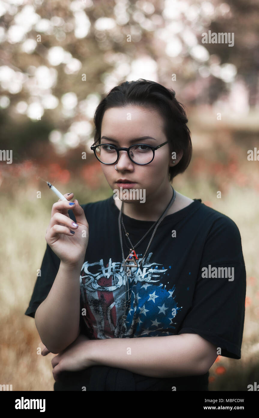 Una ragazza con corti capelli scuri, occhiali, un My Chemical Romance t-shirt e una sigaretta in un parco. Foto Stock