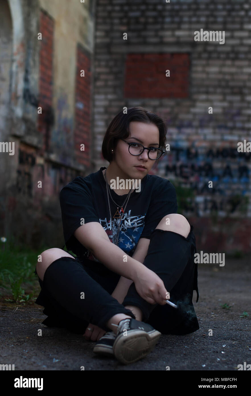 Una ragazza con corti capelli scuri, occhiali, un My Chemical Romance t-shirt e una sigaretta seduto accanto a un vecchio edificio. Foto Stock