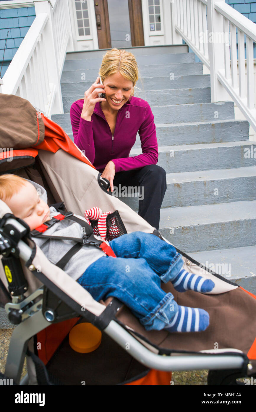 Una madre si siede sulla scalinata anteriore parlando sul suo telefono cellulare mentre un bambino dorme nella esecuzione di un passeggino. Foto Stock