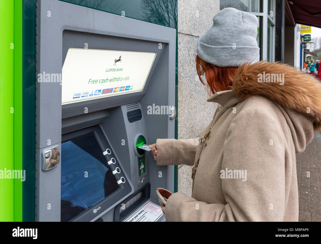 Senior donna prendendo denaro dalla Banca di Lloyds ATM in Inghilterra, Regno Unito. Sportello bancomat, punto cassa, con bancomat, il foro nella parete. Prelievo di denaro. Foto Stock