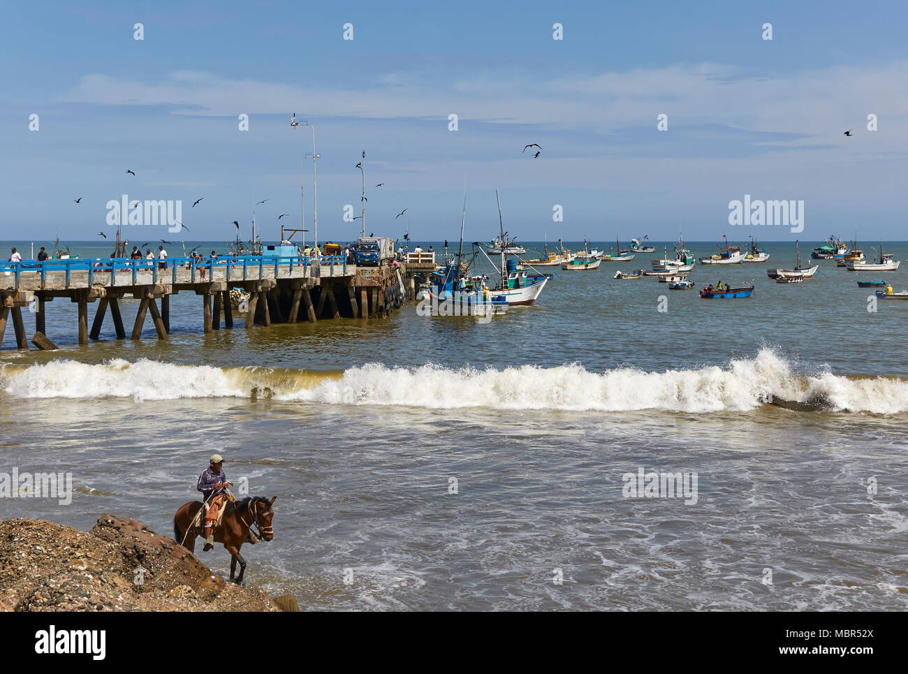I pescatori su Mancora Molo Pesca lo scarico le loro barche e supervisionare la distribuzione dei loro pesci, mentre un cavallo Cavaliere passeggiate lungo la spiaggia in F Foto Stock