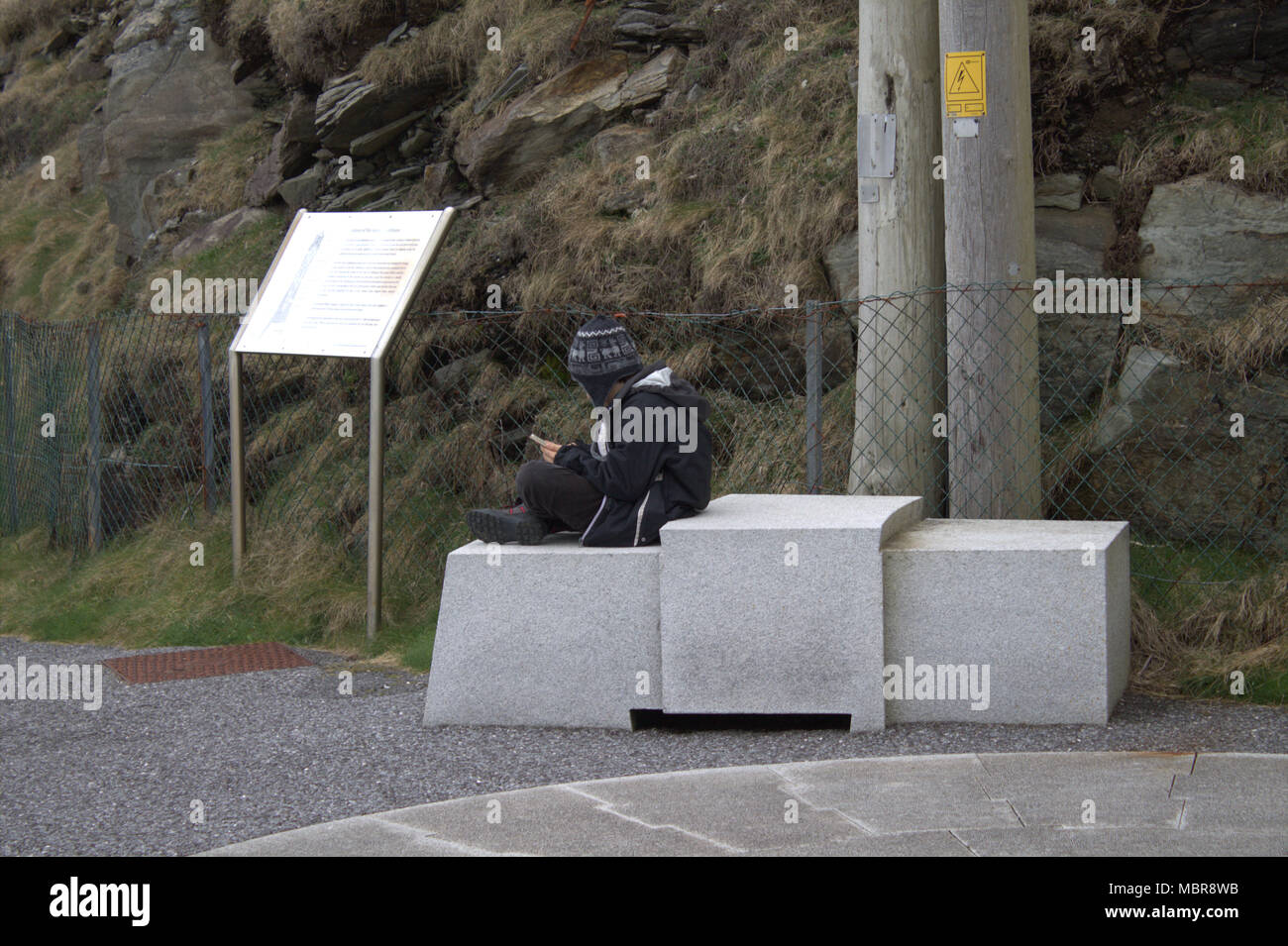 Giovane uomo seduto su lo zoccolo in pietra a Mizen Head attrazione turistica giocando sul suo telefono. Foto Stock