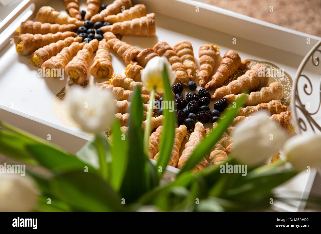 Primo piano di un buffet con un vassoio di legno con i cannoli in esso, riempito con crema pasticcera e decorata con more. Tulipani bianco sul tavolo. Foto Stock