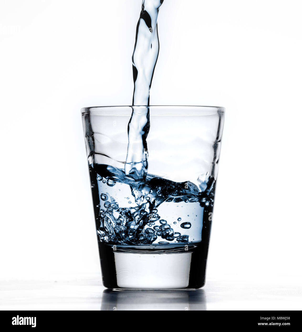 Il vetro piccolo colpo di alcool cup, essendo riempito con acqua blu, o liquido, closeup, sfondo bianco con la riflessione Foto Stock