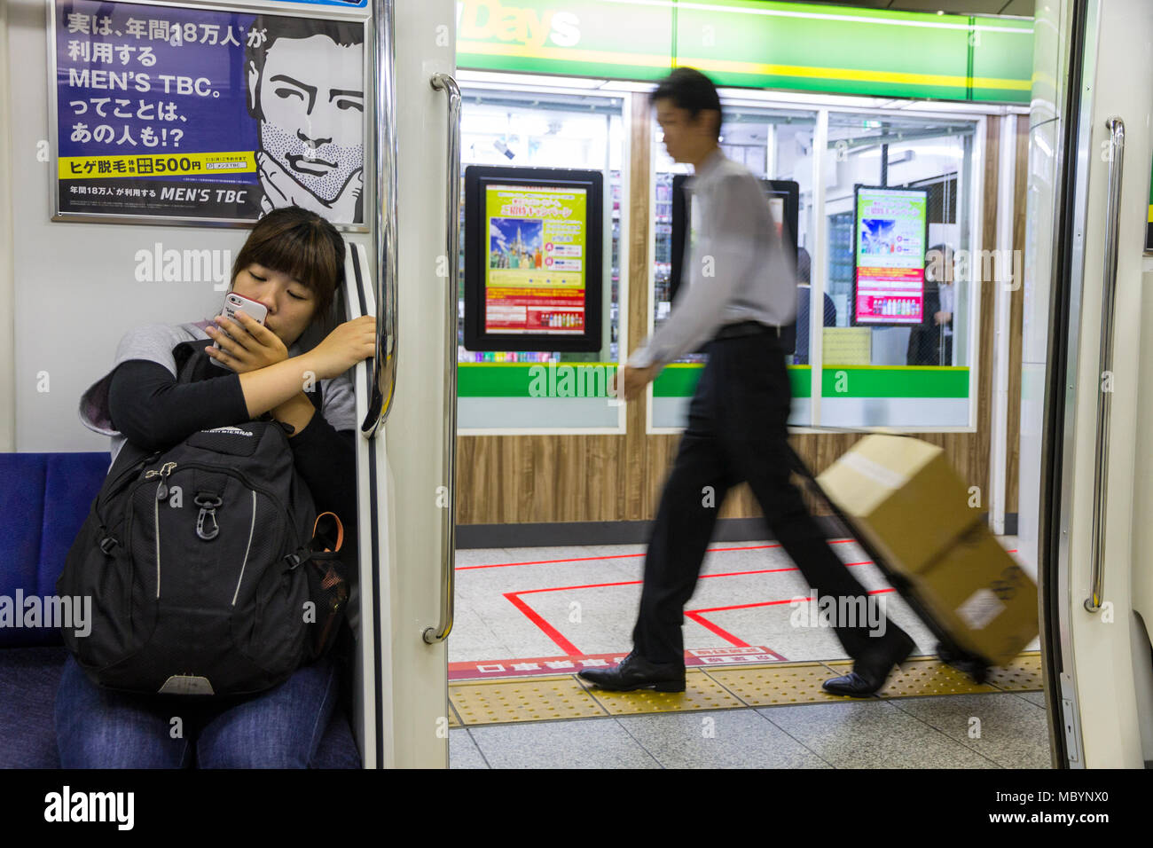 Tokyo, Giappone. Tipica scena presso la Metropolitana di Tokyo con un sit giovane ragazza guarda al suo telefono cellulare un messaggio pubblicitario, e un uomo di consegna in fretta Foto Stock