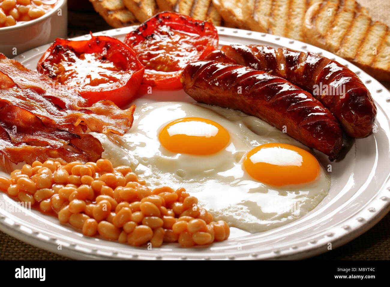 Prima colazione inglese con salsicce, pomodori grigliati, uova, pancetta, fagioli e pane sulla piastra bianca Foto Stock