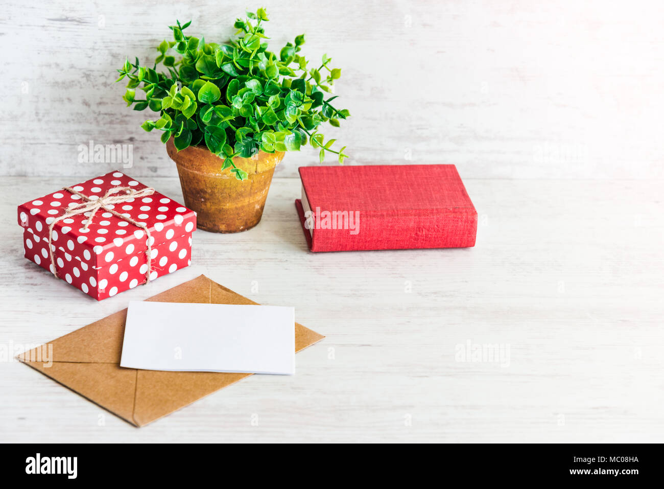 Rossa tratteggiata scatola regalo, scheda vuota, kraft busta, libro rosso e verde fiore in una rustica vaso di ceramica. Bianco sullo sfondo di legno, copia dello spazio. Foto Stock