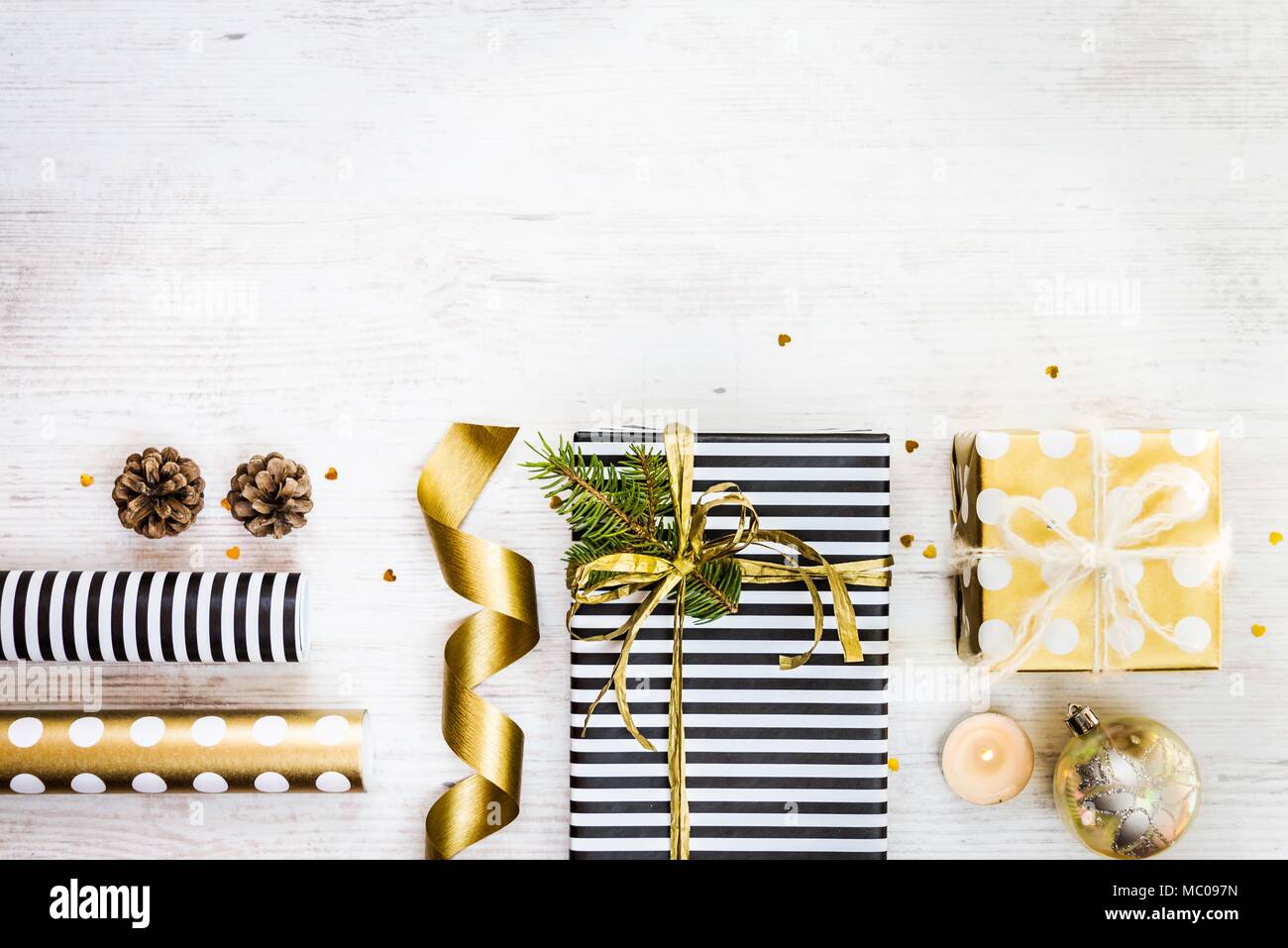 Confezioni regalo avvolto in bianco e nero a strisce e dorata punteggiata con carta, pino, coni, candela e i materiali di confezionamento su un legno bianco sfondo vecchio. Foto Stock