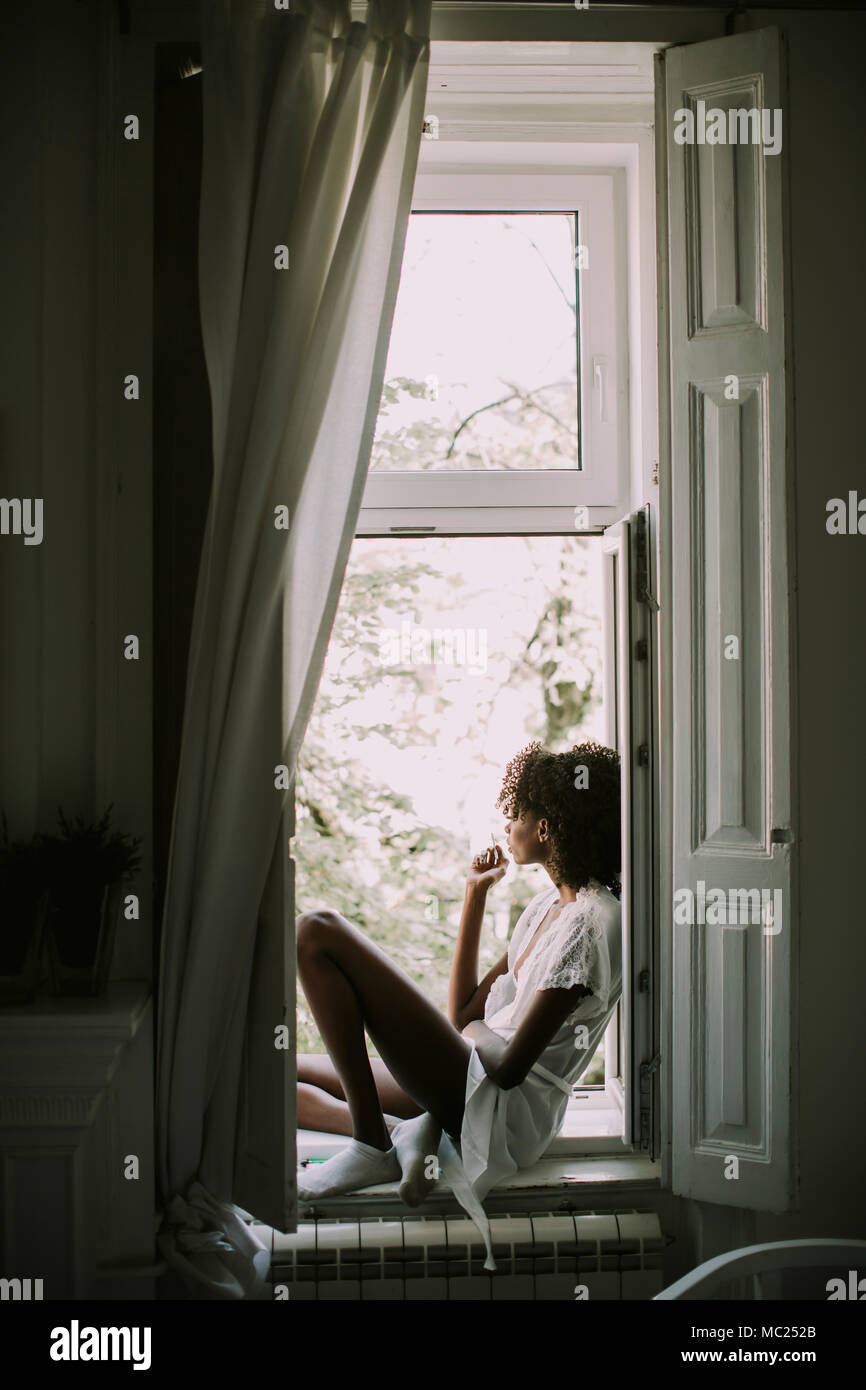 Piuttosto giovane nero donna seduta sulla finestra in camera e Sigaretta fumare Foto Stock