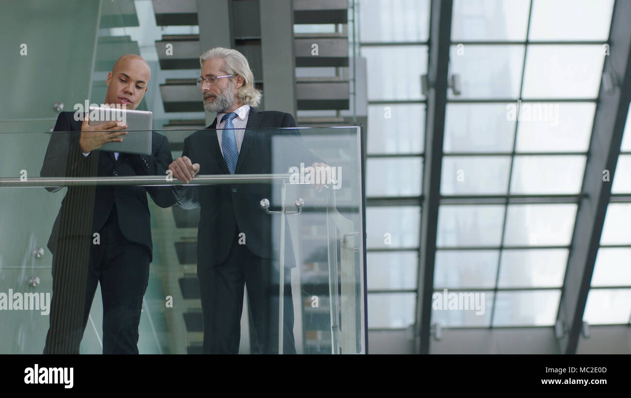 Latino e caucasico corporate executives in piedi sul secondo piano di un edificio moderno a discutere di business utilizzando digitale compressa. Foto Stock