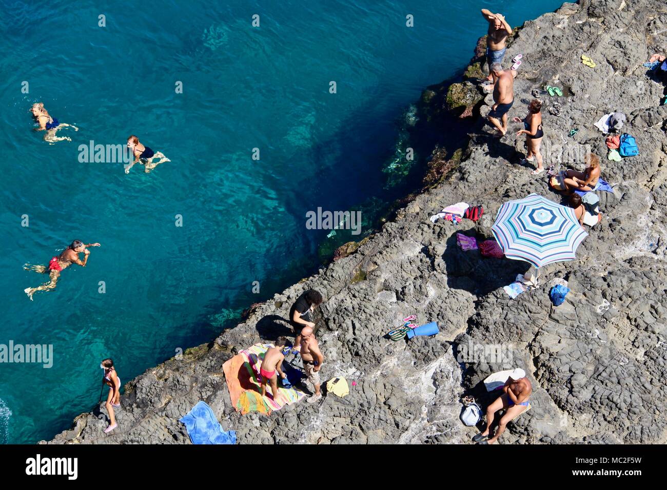 Una veduta aerea di Aci Castello di rocce al di sotto vicino le acque blu tropicali a Catania, in Sicilia. Persone in abiti nuotare e ombrelloni in un caldo giorno d'estate. Foto Stock