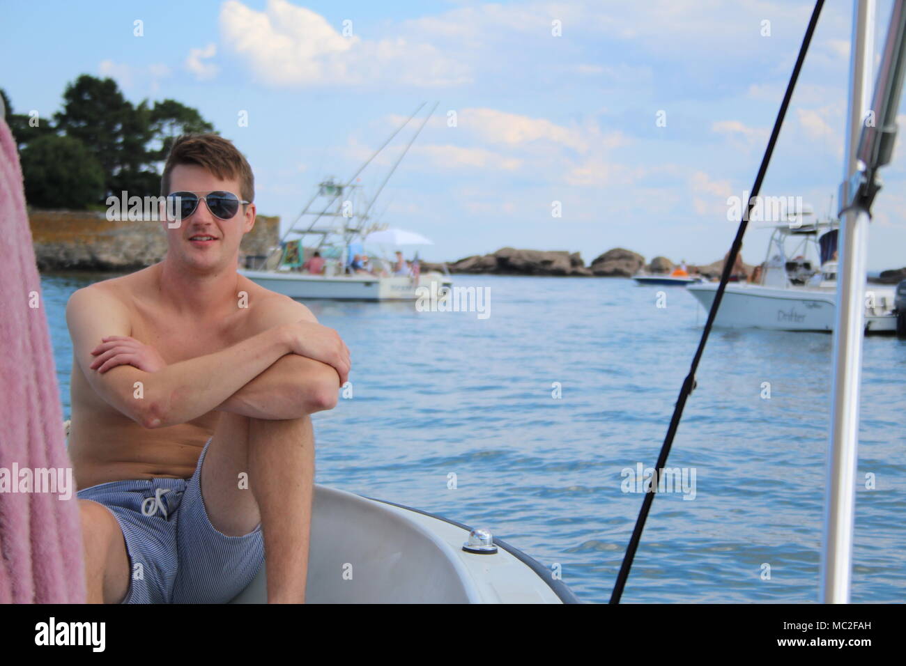 Un uomo con gli occhiali da sole e blu pantaloncini spelati su una barca in Rockport. Estate di relax con amici e barche in background. Foto Stock