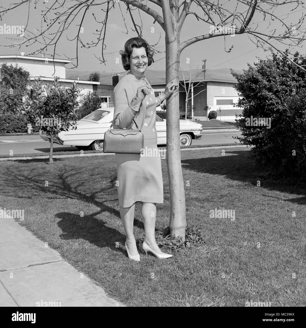 Una donna felice poggia su una struttura ad albero e tenere la sua borsa la California in un quartiere residenziale, ca. 1965. Foto Stock