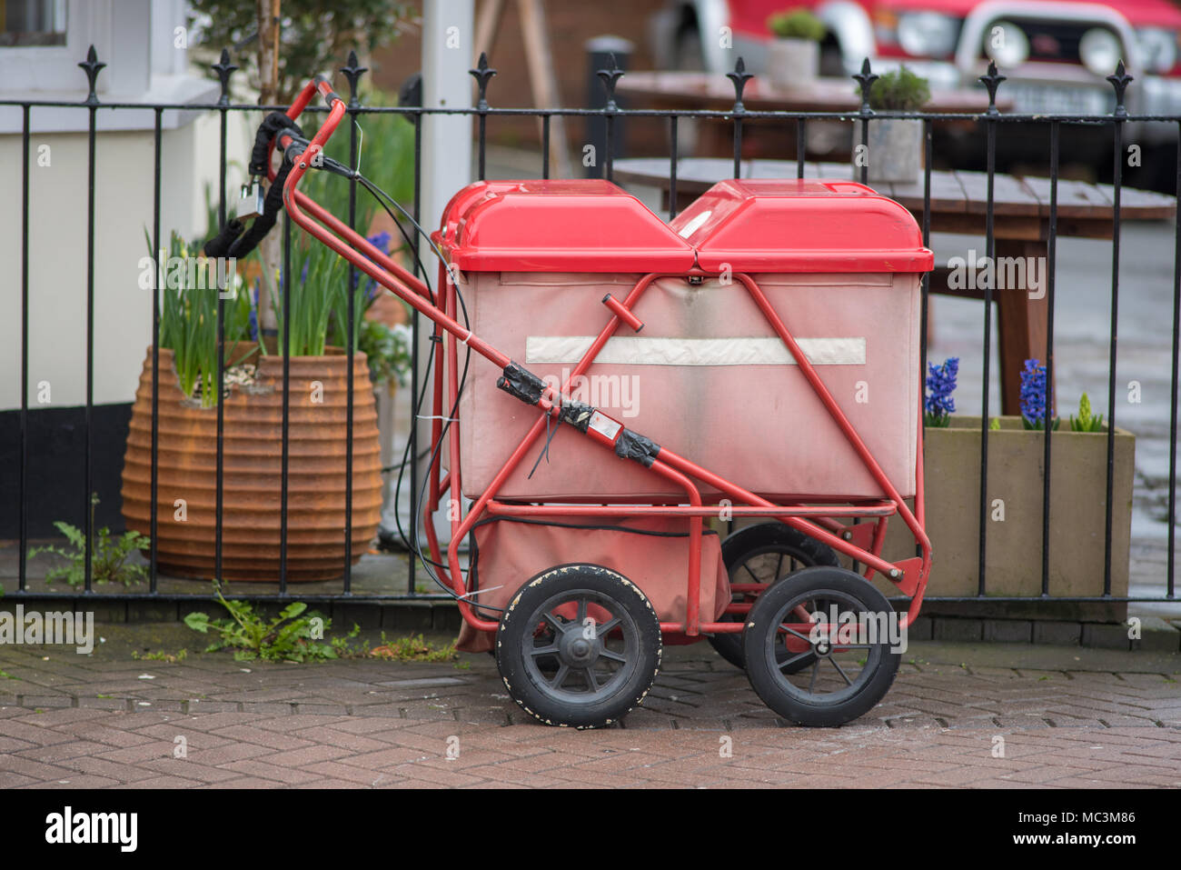 Rosso brillante royal mail spingere il carrello barrow incatenato al cast recinzione in ferro con giardino in background Foto Stock