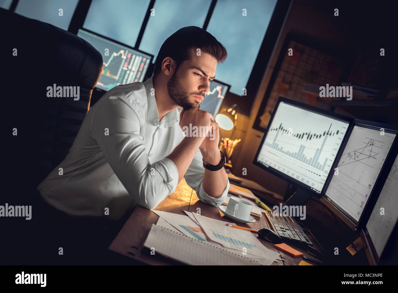 Concentrato considerato trader o investitore serio lavoro notturno le ore di lavoro straordinario analizzando le negoziazioni di borsa grafici guardando i monitor dei computer controllo di mercato Foto Stock