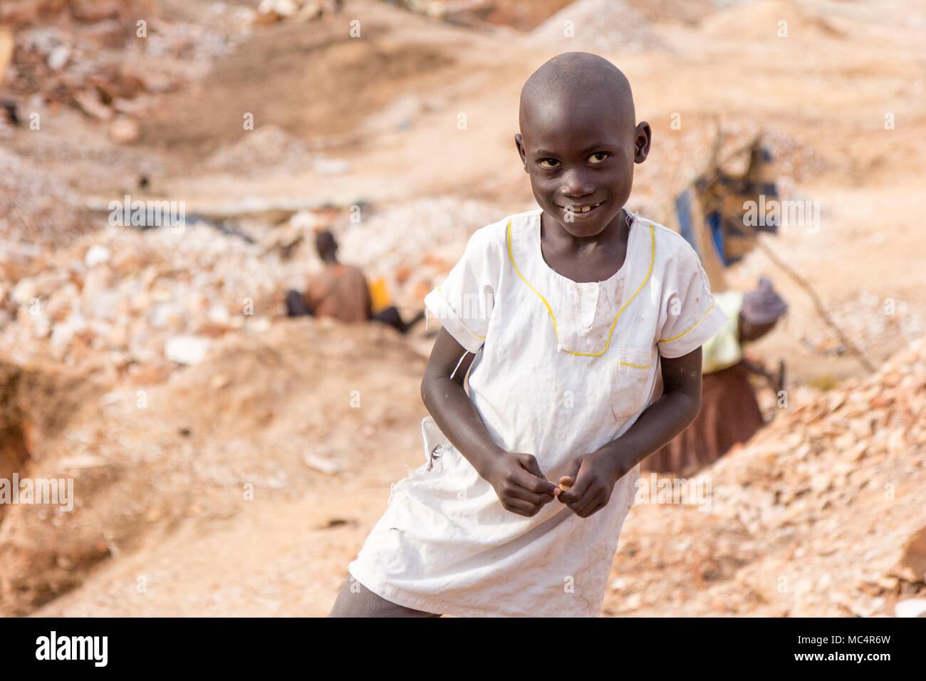 Lugazi, Uganda. Il 18 giugno 2017. Un sorridente ragazzo ugandesi in piedi in una cava. Persone in background sono rocce di rottura in lastre di piccole dimensioni per la vendita al Foto Stock