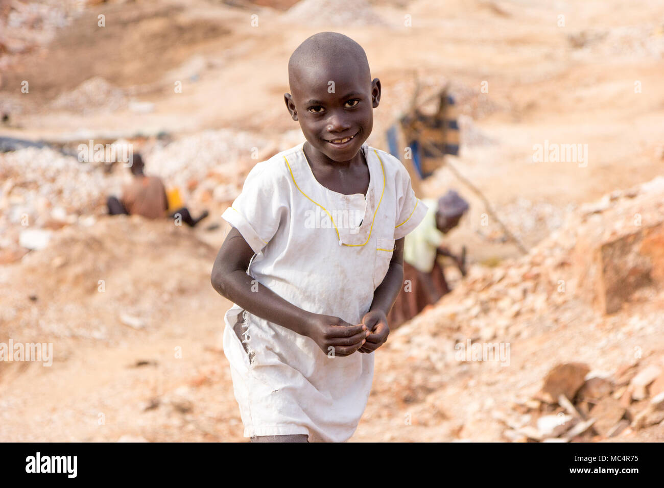 Lugazi, Uganda. Il 18 giugno 2017. Un sorridente ragazzo ugandesi in piedi in una cava. Persone in background sono rocce di rottura in lastre di piccole dimensioni per la vendita al Foto Stock