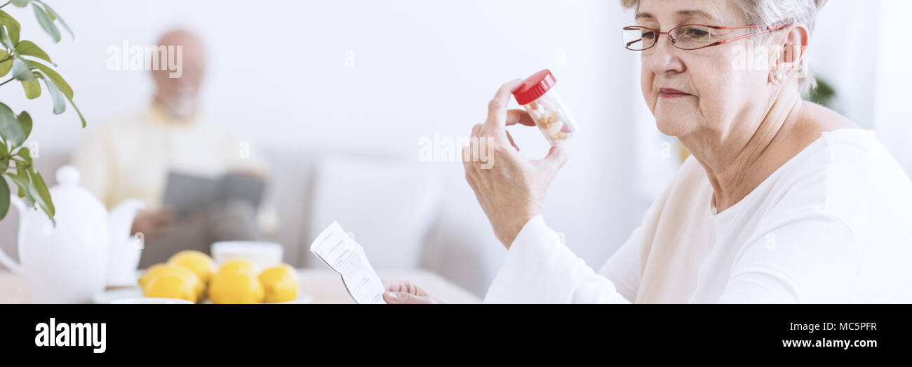 Un senior donna che indossa gli occhiali guardando una bottiglia trasparente con le pillole e tenendo un farmaco foglietto informativo. Un sfocata la figura di un uomo più anziano di leggere Foto Stock