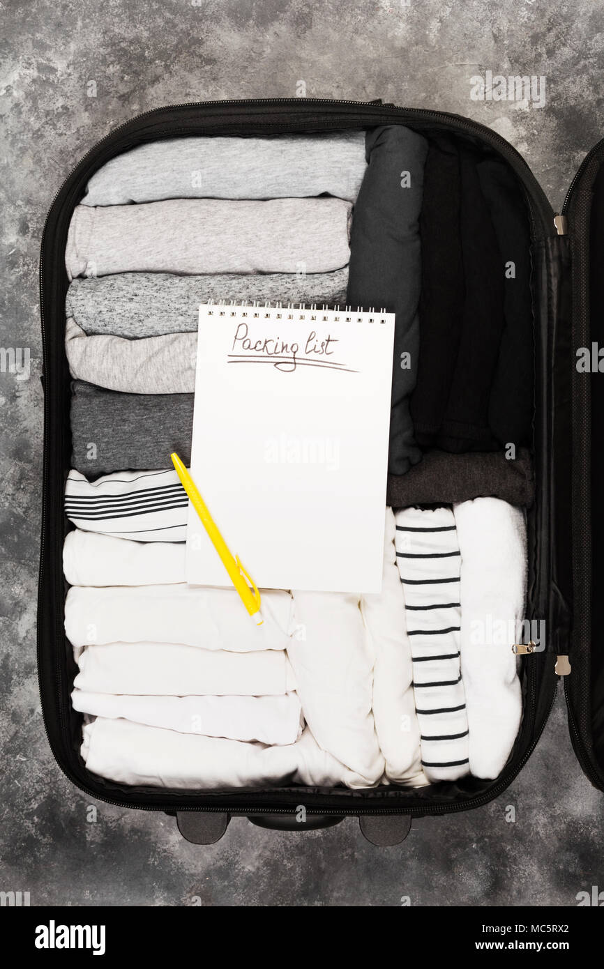 Confezionamento di abiti monocromatici in bianco e nero la valigia. Lista di imballaggio in bianco per notebook. Vista superiore, spazio di copia Foto Stock