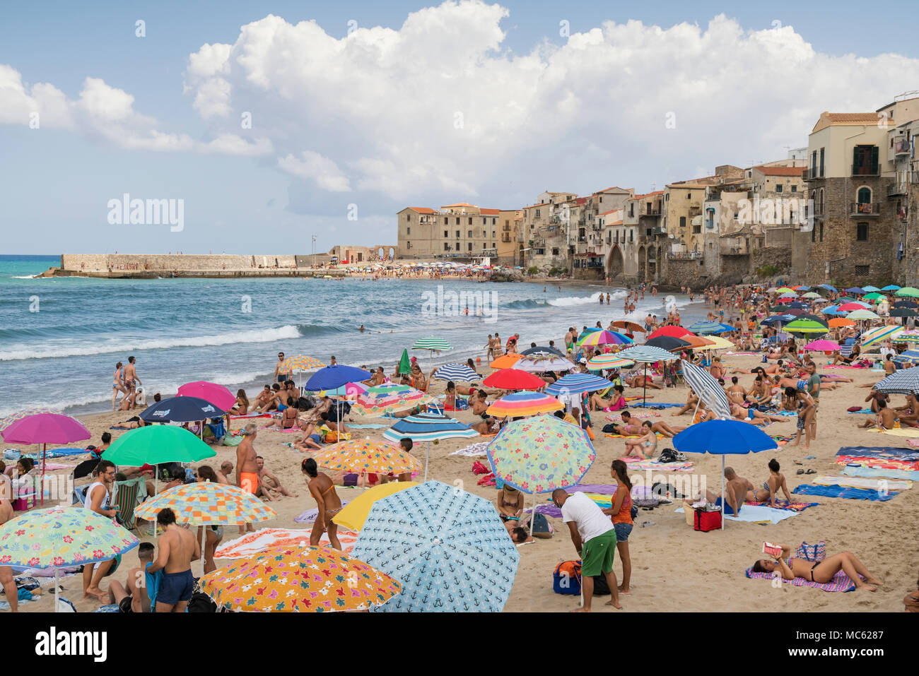 Cefalù, Sicilia, Italia, una folla di gente che coprono la spiaggia sabbiosa godendo le vacanze su un luminoso giorno di agosto con la città vecchia di edifici come un bac Foto Stock