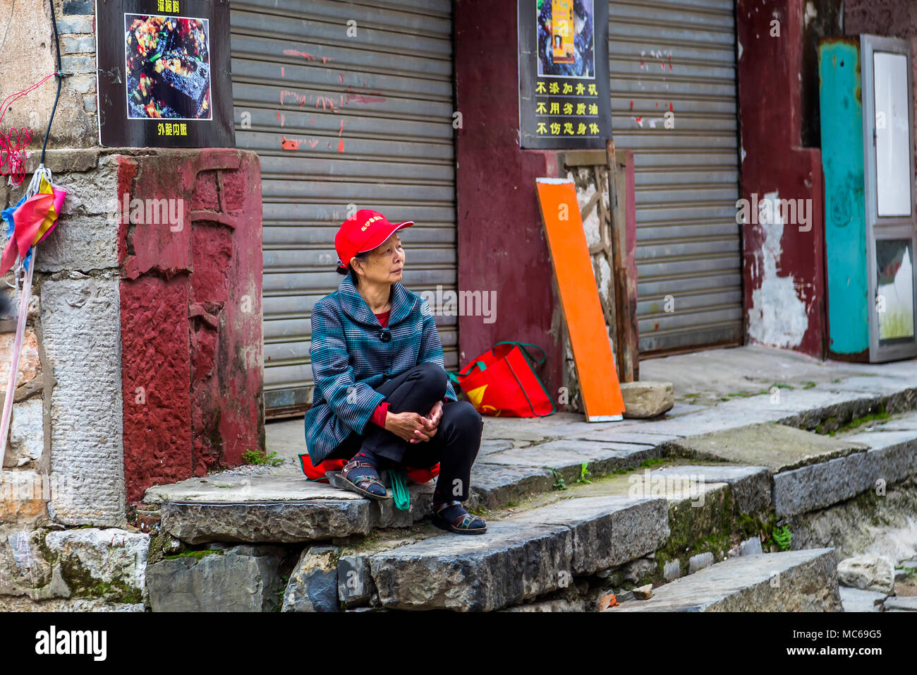 Un anziana signora cinese guarda pensieroso come lei siede su alcuni gradini in pietra nel borgo antico di Daxu, Cina. Foto Stock