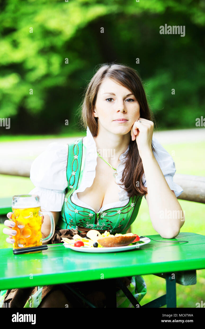 Giovane donna bavarese in dirndl seduta a tavola nel giardino della birra con birra e cibo Foto Stock