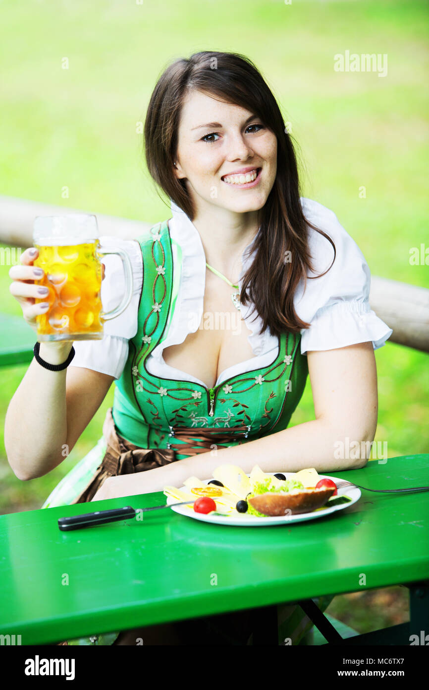 Giovane donna bavarese in dirndl seduta a tavola nel giardino della birra con birra e cibo Foto Stock