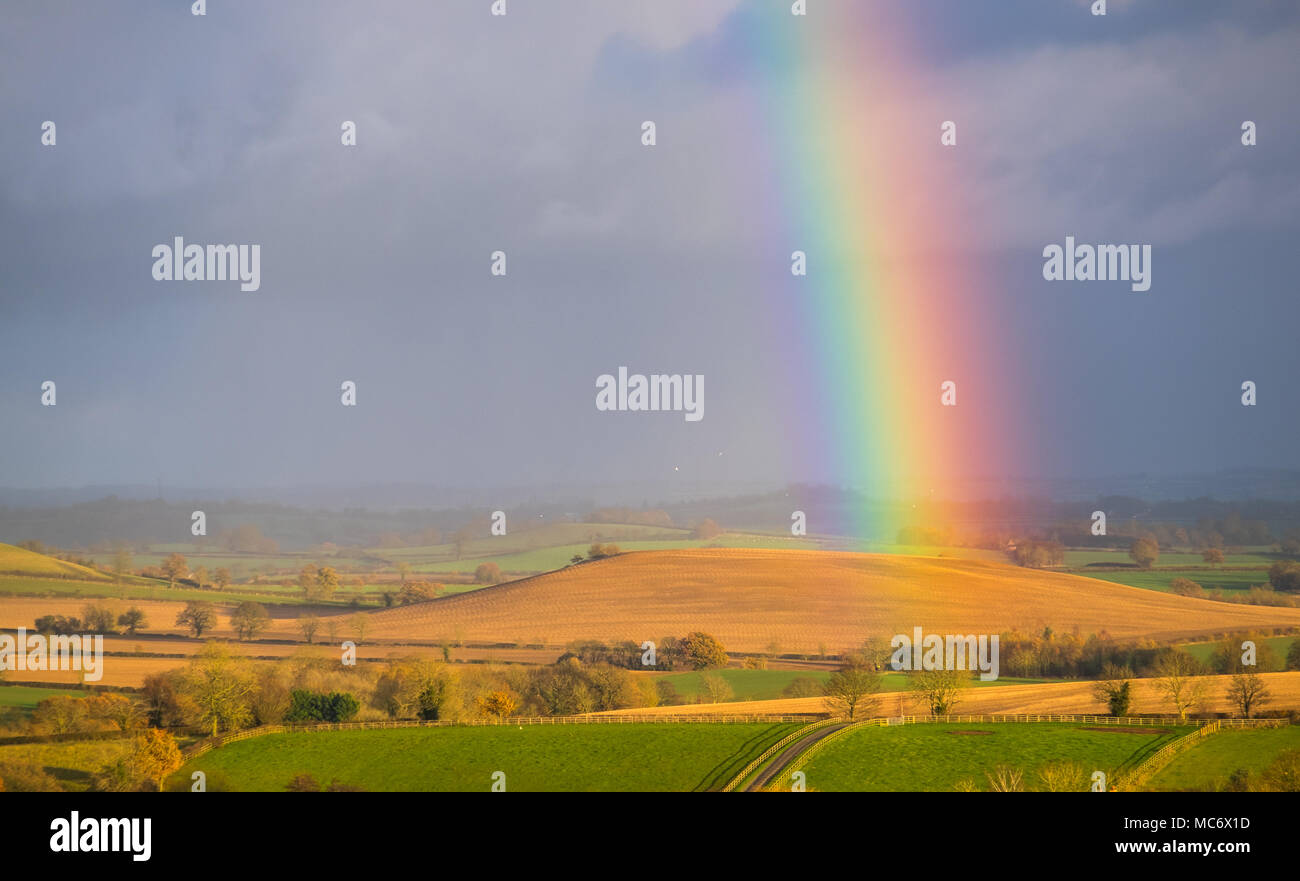 Vivacemente colorato rainbow contro il grigio scuro cielo, sulle ondulate colline dorate in nord Oxfordshire nella luce di abbassamento. Foto Stock