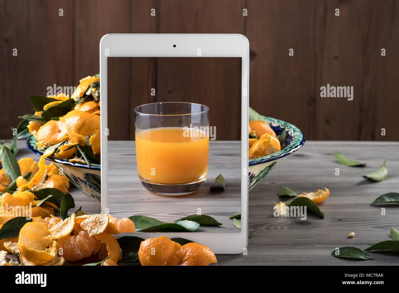 Bianco tablet mobili in piedi nella parte anteriore del piatto di ceramica riempito tangerini freschi grigio su un tavolo di legno. Pop up effetto visivo. Creative composizione alimentare. Foto Stock