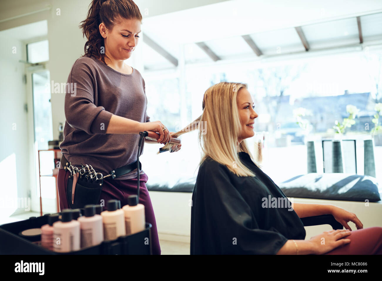 Sorridente bionda e giovane donna seduta in un salone sedia avente i suoi capelli raddrizzato dal suo parrucchiere Foto Stock