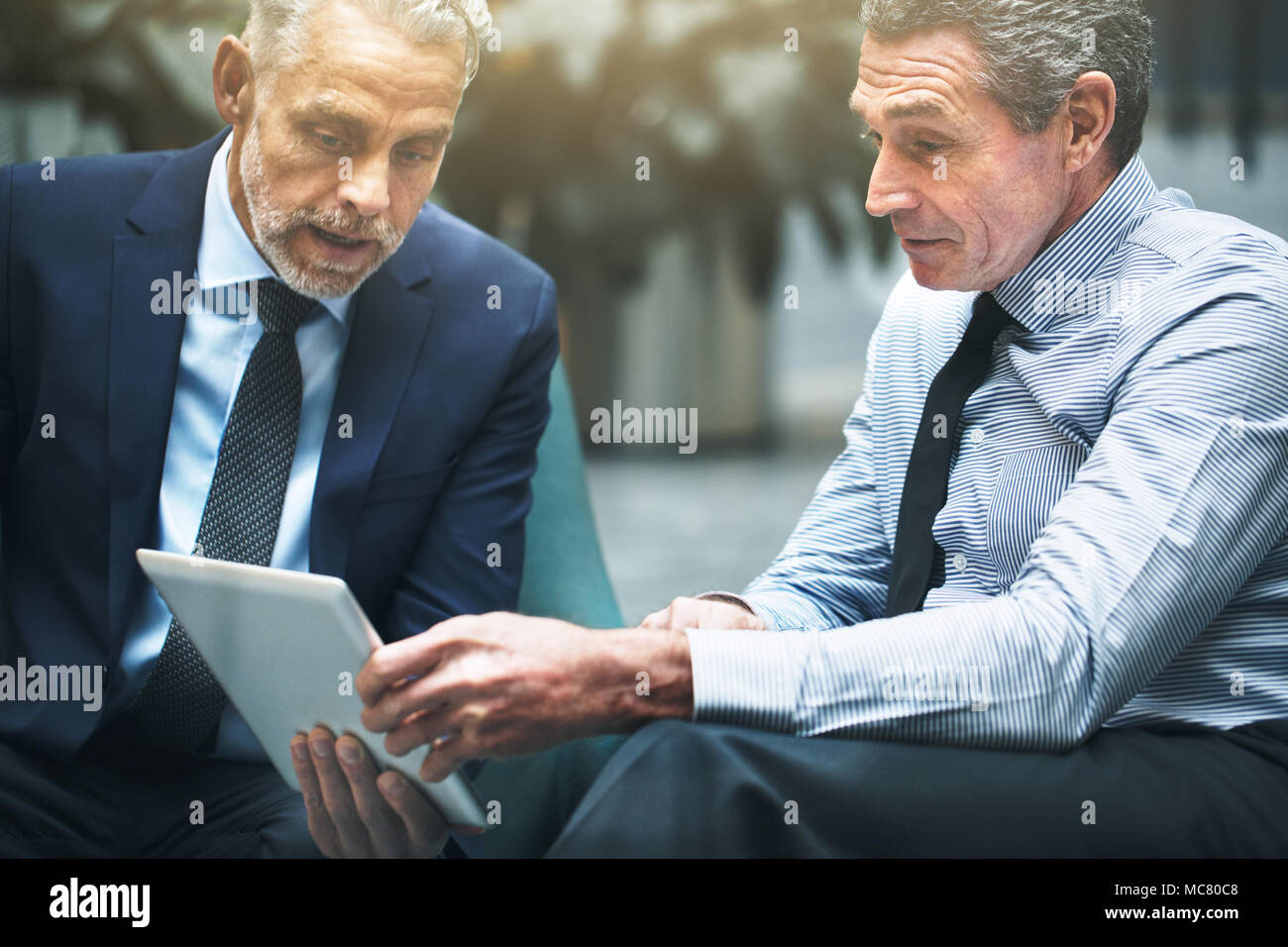 Due ben vestito imprenditori maturi a discutere di lavorare insieme e utilizzando una tavoletta digitale seduti su sedie in un ufficio lobby Foto Stock
