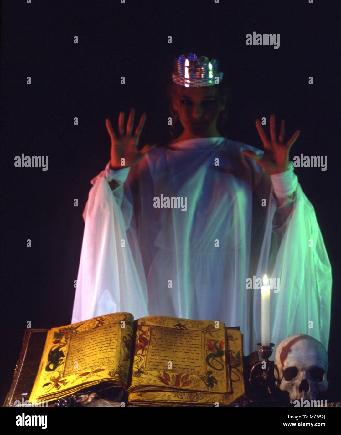 La stregoneria moderna strega, indossando rituale bianche vesti, in gront di un altare con un grimoire, cranio, rituale spada, candela etc Foto Stock