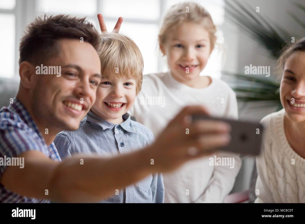Kid sister rendendo poco carino fratello bunny orecchie mentre padre tenendo la famiglia felice selfie, funny bambini un ragazzo e una ragazza divertirsi in posa per self-portr Foto Stock