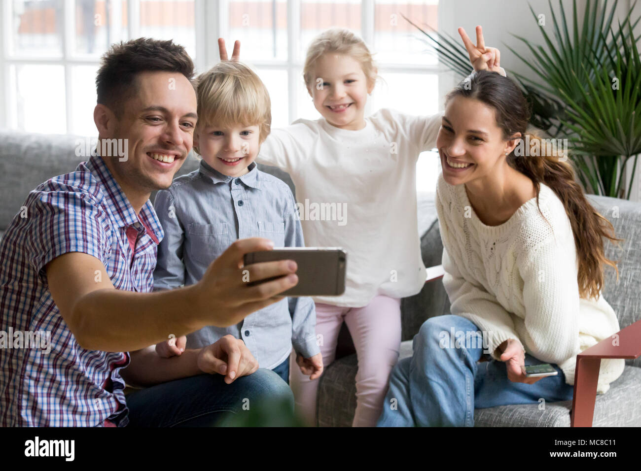 Ragazza sorridente rendendo la madre e il fratello bunny orecchie mentre padre tenendo la famiglia felice selfie, giocoso i bambini adottati avendo divertimento che posano per una foto divertente Foto Stock