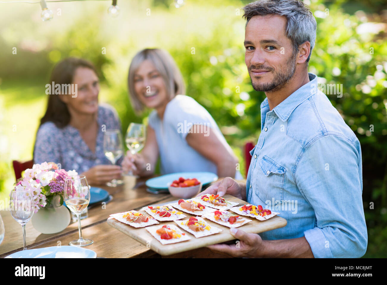 Gruppo di amici in quarantenni si sono riuniti intorno ad un tavolo in giardino per condividere un pasto. Un uomo offre spuntini per gli ospiti mentre guardando la telecamera Foto Stock