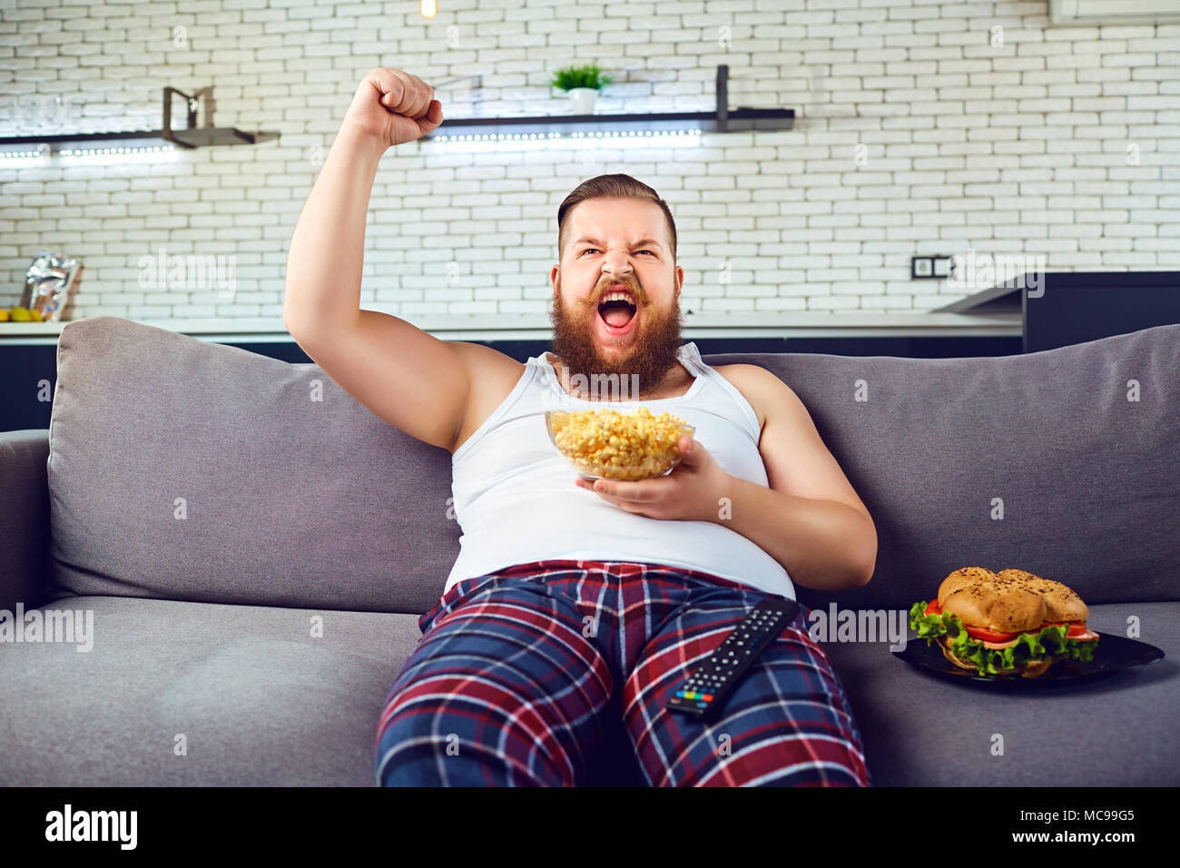 Spessore funny man in pigiama mangiare un hamburger seduti sul divano guardando la TV a casa. Foto Stock