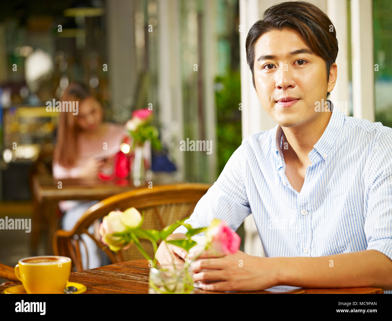 Giovane uomo asiatico seduti ad un tavolo nel negozio di caffè o di tè casa con una giovane donna in background. Foto Stock