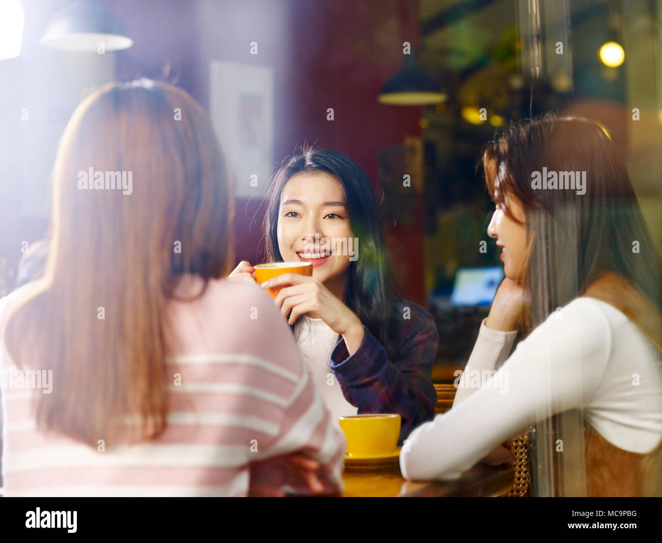 Tre felice giovane e belle donne asiatiche seduta a tavola in chat parlando in negozio di caffè o tè casa, shot attraverso il vetro di una finestra. Foto Stock