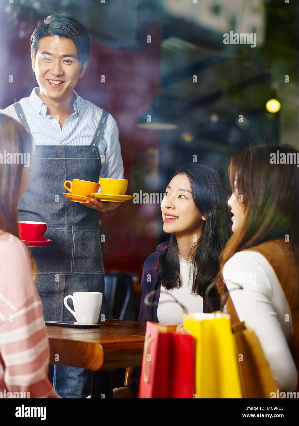 Giovani sorridenti asian cameriere che serve i clienti di sesso femminile nel coffee shop, shot attraverso il vetro di una finestra. Foto Stock