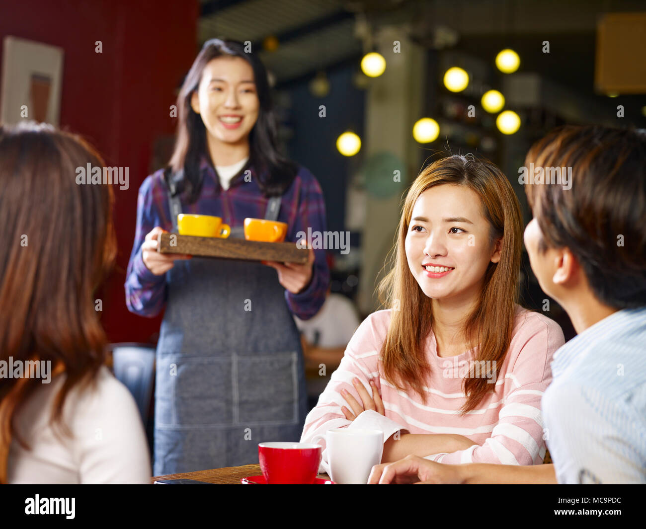 Tre felice giovani asiatici adulti seduti a tavola in chat a parlare mentre la cameriera che serve caffè, concentrarsi sulla ragazza in primo piano. Foto Stock