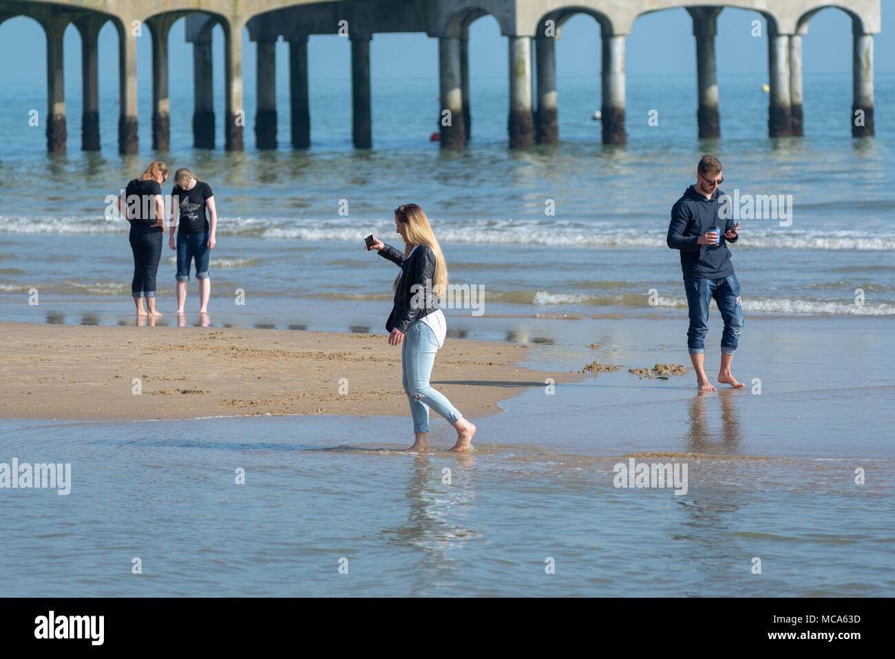 La gente scarta scarpe e calzini e pagaia in mare con pantaloni arrotolati. Passeggiate sulla spiaggia in una calda giornata di aprile, Boscombe, Bournemouth, Dorset, Regno Unito Foto Stock