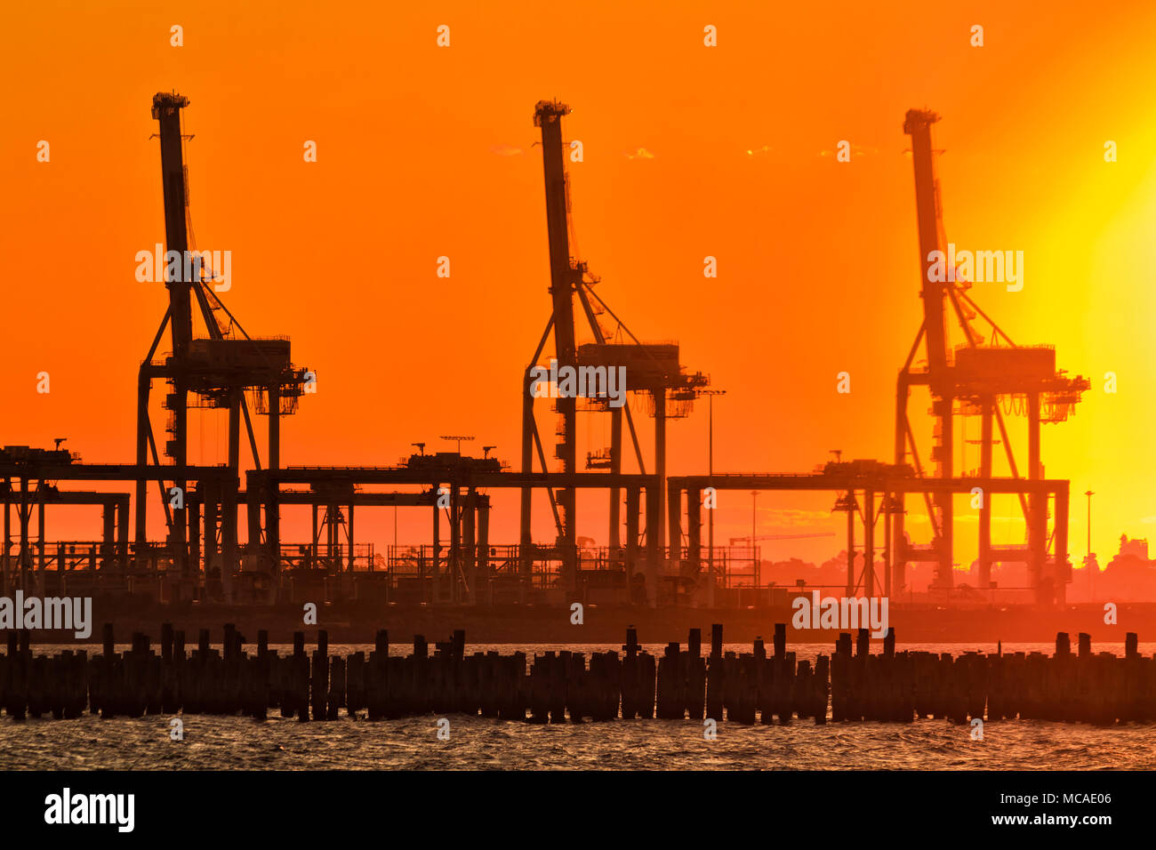 Rosso arancione bruciato il cielo al tramonto contro l'impostazione caldo sole alto contrasto pesanti carichi industriali gru da carico in Port Melbourne della Baia di Port Phillip Foto Stock