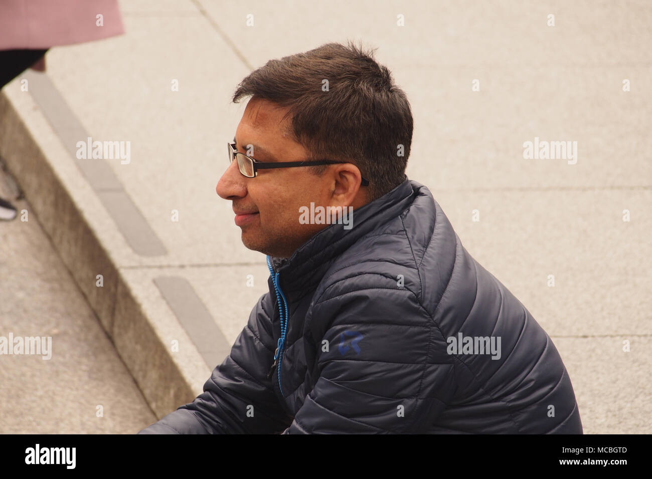 Un giovane uomo seduto sui gradini di pietra in Trafalgar, Londra a guardare il mondo che passa da indossare una folta pelliccia e bicchieri Foto Stock