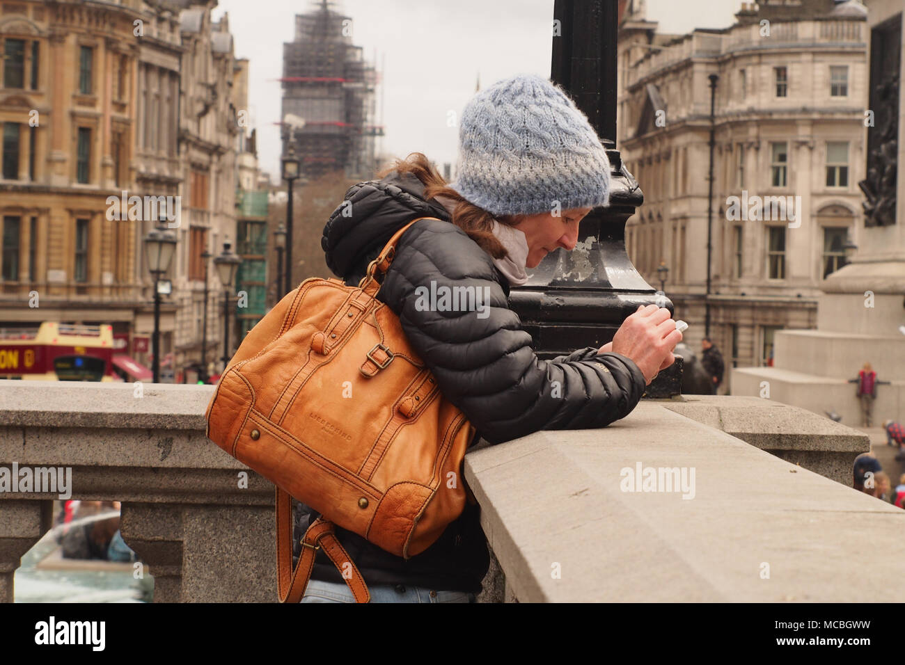Una donna che indossa un cappotto di spessore, cappellino e borsa, poggiando su una parete in Trafalgar Square, Londra guardando verso il basso e concentrandosi sullo smartphone Foto Stock