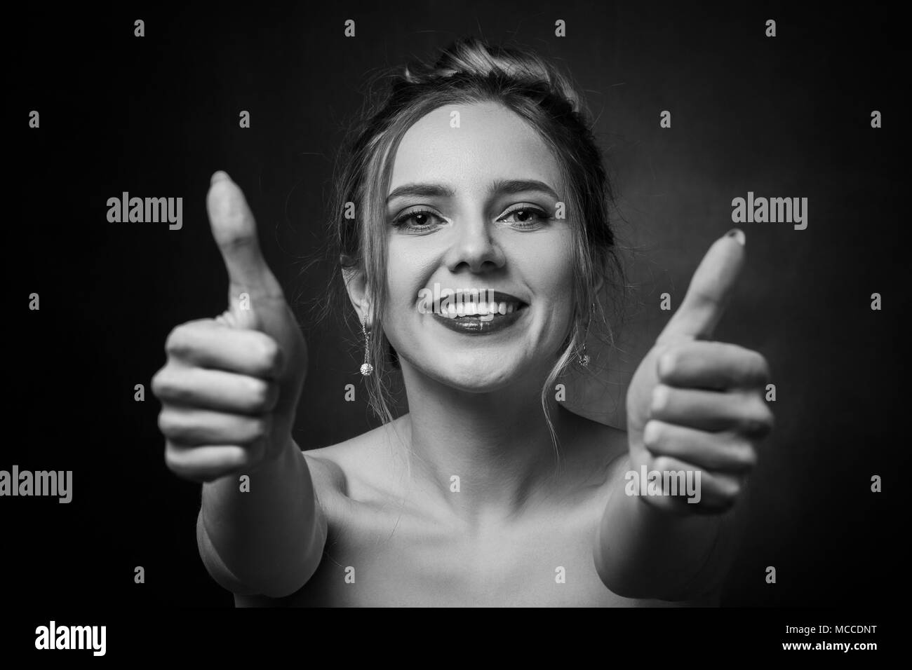 Donna felice mostra il suo pollice su sfondo nero guardando la telecamera ridendo, monocromatico Foto Stock