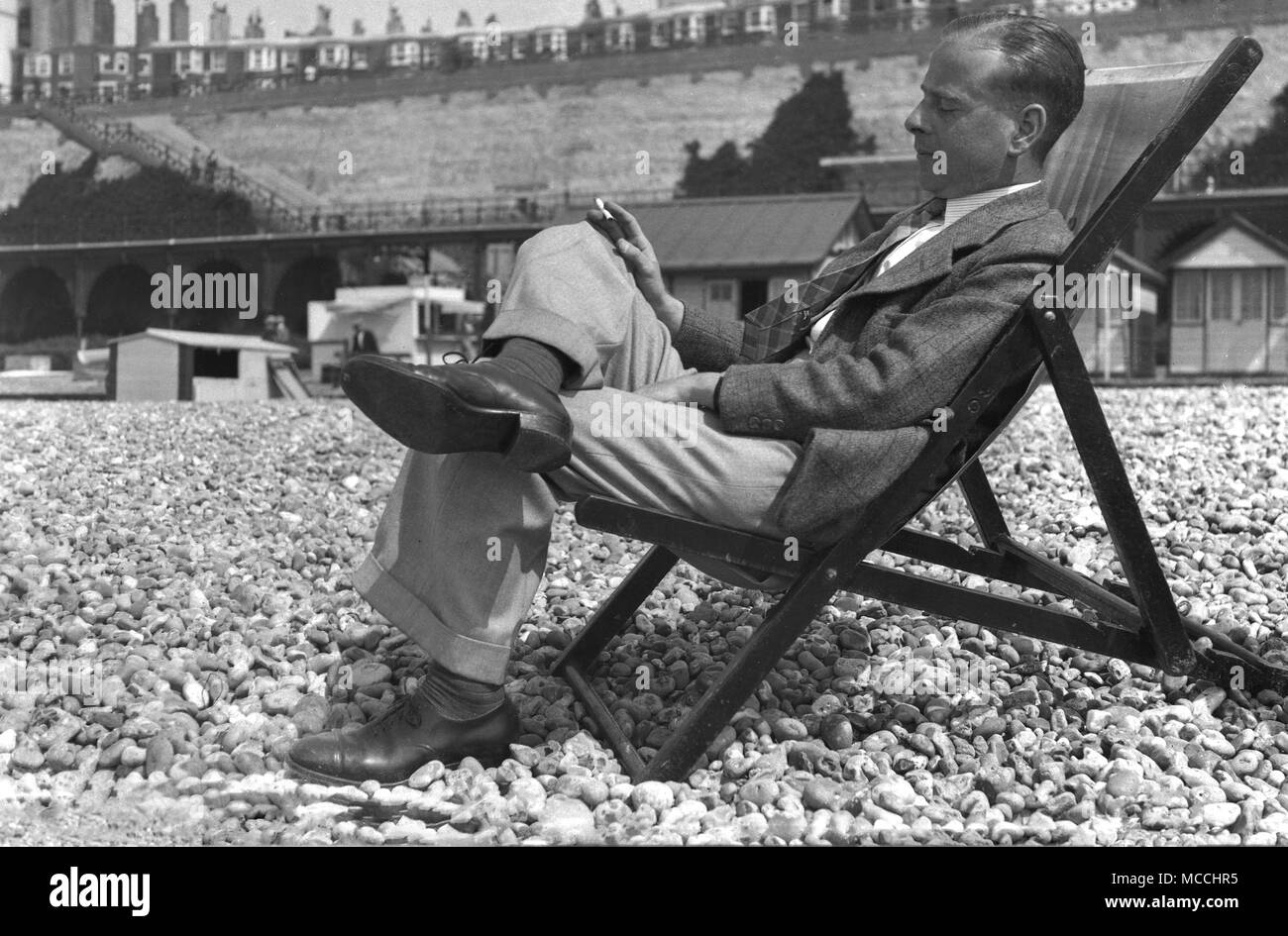 Fine degli anni quaranta, storico, un ben vestito gentleman godendo di una sigaretta e un pisolino - un po' di shut-eye - mentre vi rilassate sulla propria seduto in una sedia a sdraio al di fuori su di una spiaggia di ciottoli, Inghilterra, Regno Unito. Foto Stock