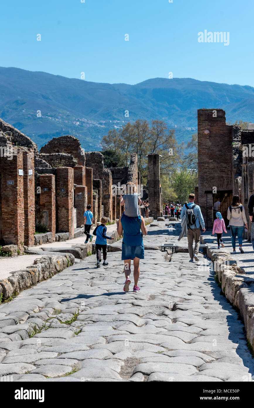 Kids in Pompei esplorare una strada rocciosa con resti di strutture e di colline in background con la famiglia, un genitore mamma trasporta un bambino piccolo sulle spalle Foto Stock