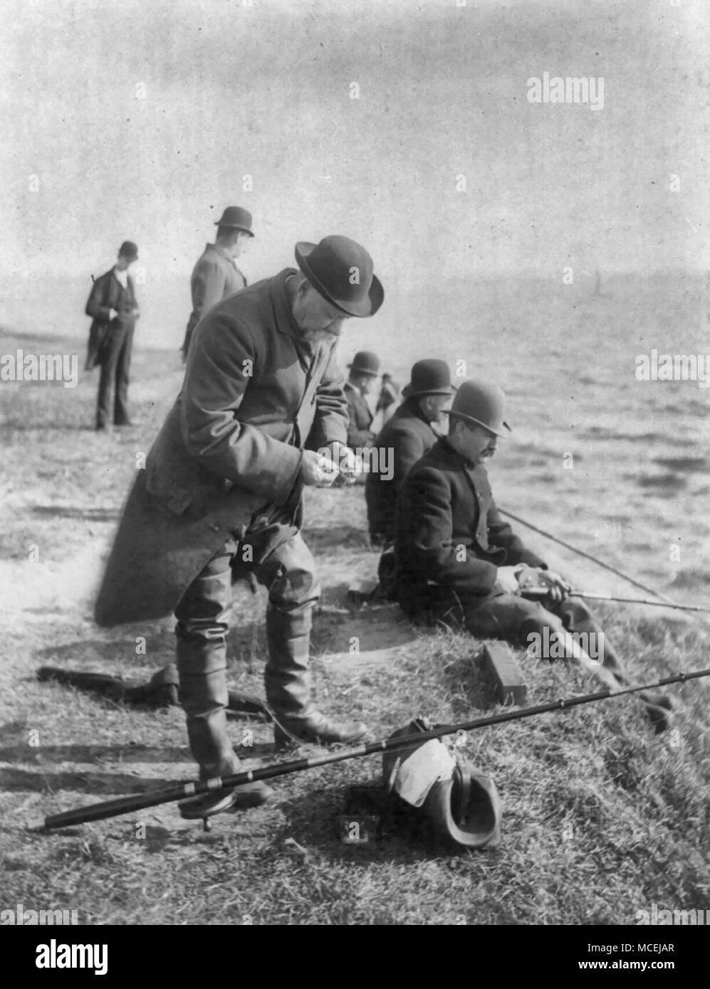 Un veterano pescatore - Uomo hook baiting indossando swallow-tailed cappotto e altri uomini seduti sulla banca e la pesca nelle Montagne Adirondack, N.Y., circa 1890 Foto Stock